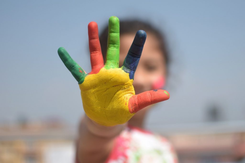 child's hand each finger painted in green, yellow, green, blue and red with child face blurred in the background